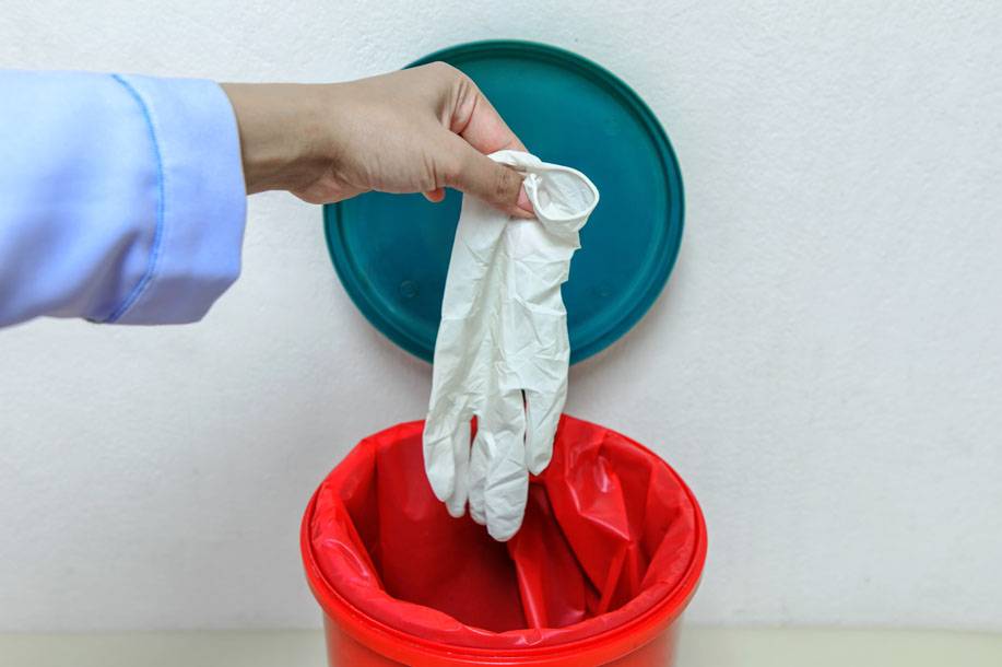 A hand drops a used glove into a medical waste bin.